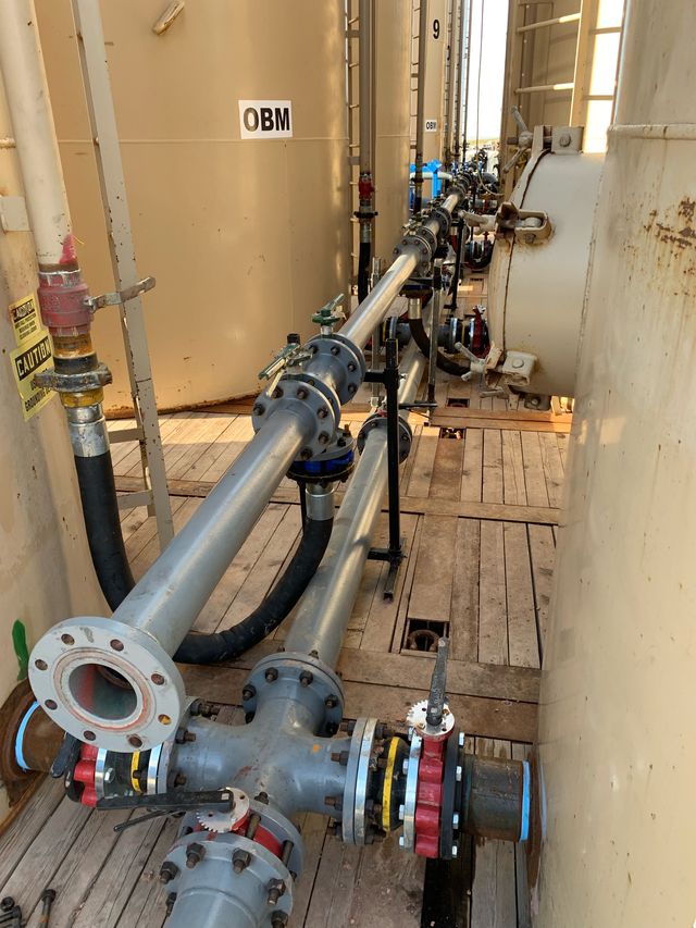 Long run of gray flanged pipe spools along a wooden walkway between tanks, with a cross fitting and red and blue butterfly valves in the foreground