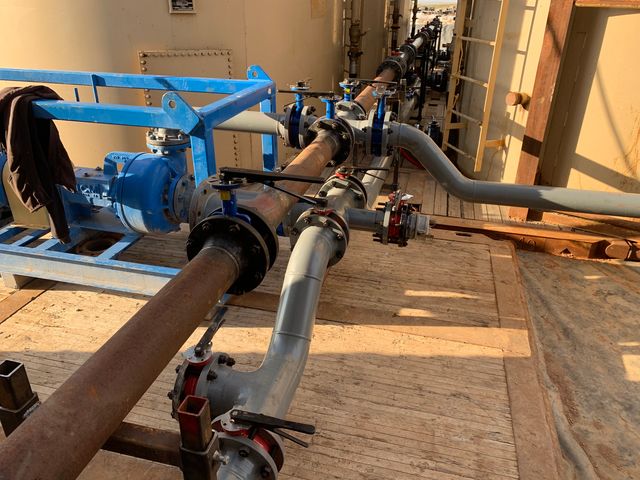 Wide view of a valve manifold and gray pipe spools tied into a pump on a blue skid, with the pipe run continuing along the tanks