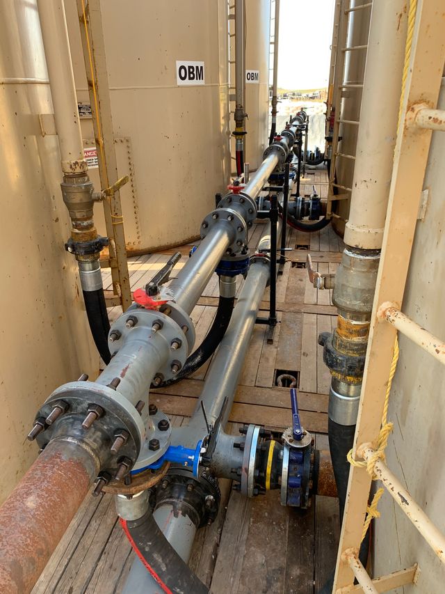 Flanged pipe run with valves and hoses along a narrow wooden walkway between tanks