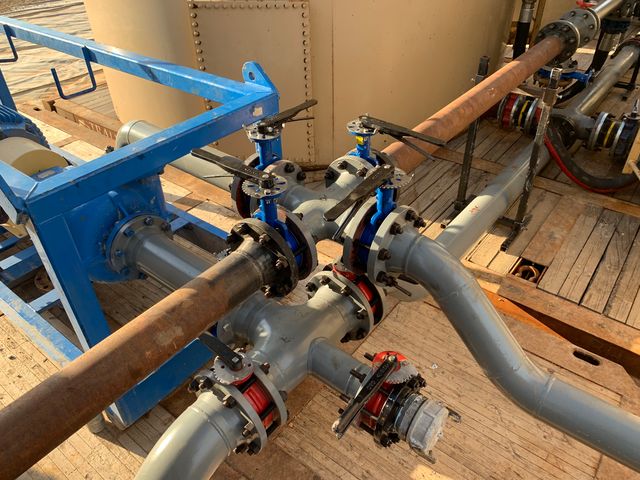 Valve manifold with blue and red butterfly valves on gray flanged pipe, next to a blue pump skid