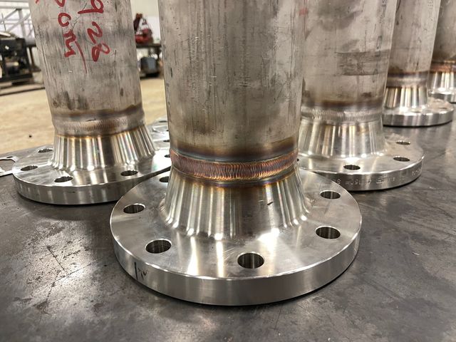 Close-up of a TIG weld joining stainless steel pipe to a weld-neck flange, with more stainless spools on the shop table behind it