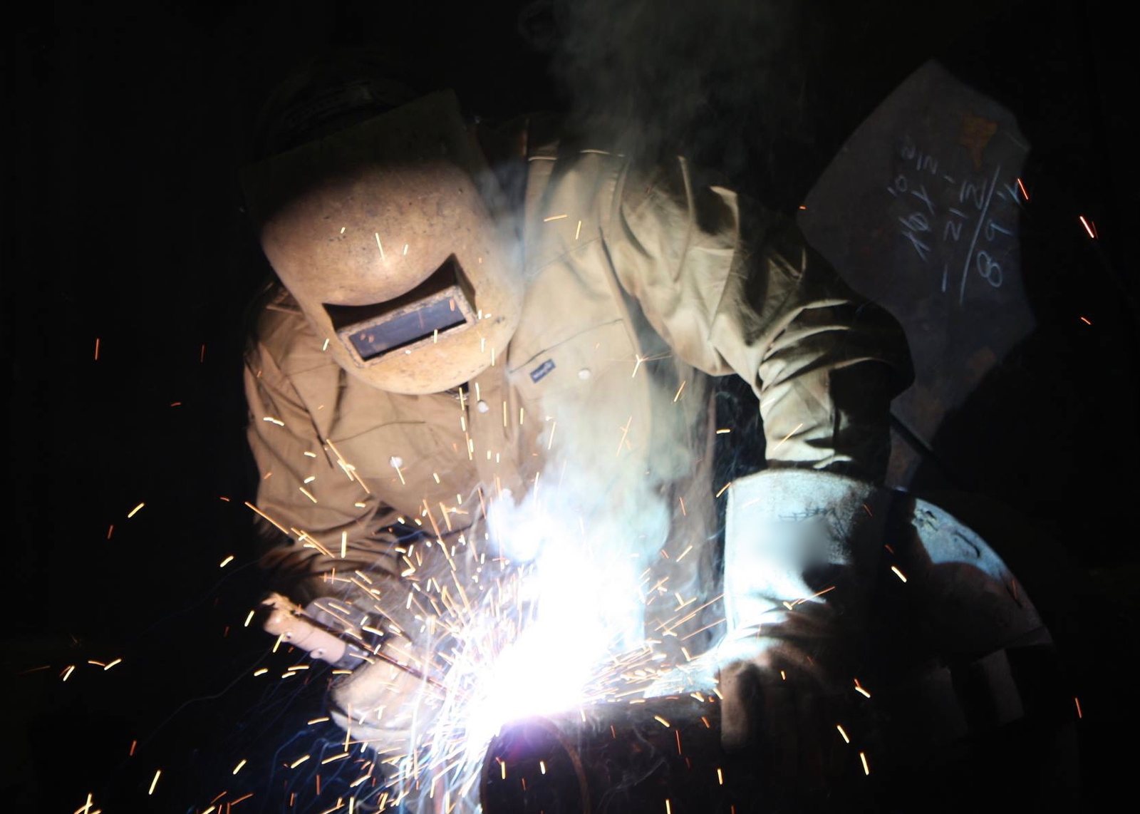 Welder in a welding hood running a weld, with sparks flying against a dark background