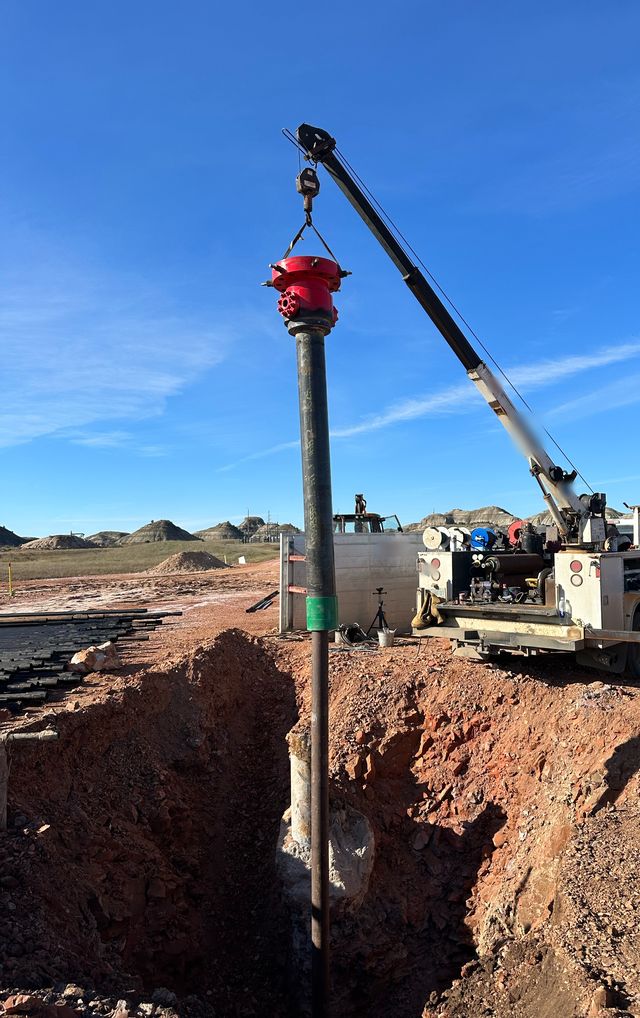 Red wellhead hanging from a service truck crane over a pipe in an excavated well site, under a blue sky