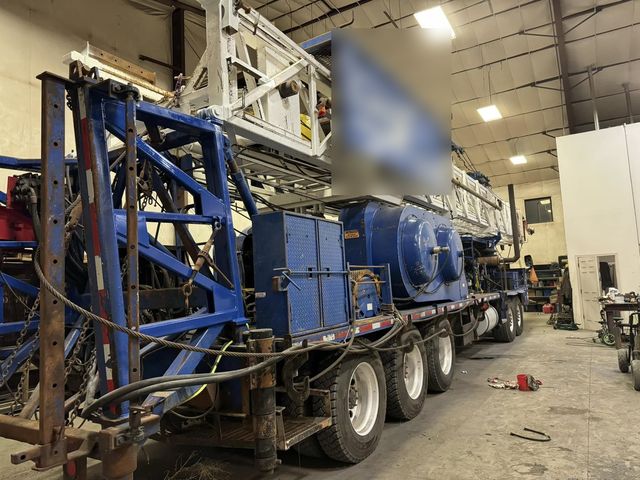 Blue production (workover) rig with its derrick lowered, inside the HWF shop for repair